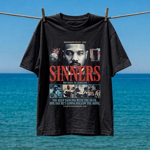 May include: A black short-sleeve t-shirt featuring a graphic print for the film Sinners. The design includes a portrait of Michael B. Jordan, the text MISSISSIPPI DELTA - 1932, SINNERS, and the quote YOU KEEP DANCING WITH THE DEVIL, ONE DAY HE'S GONNA FOLLOW YOU HOME.