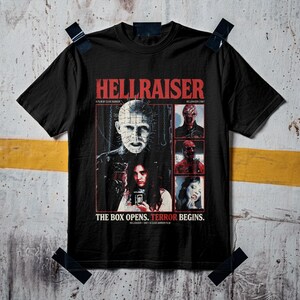 Może przedstawiać: Czarny t-shirt z nadrukiem graficznym z filmu Hellraiser. Projekt zawiera tekst HELLRAISER, THE BOX OPENS. TERROR BEGINS. oraz obrazy Pinheada i innych postaci z filmu z 1987 roku, ułożone w prostokątnym układzie na środku koszulki.