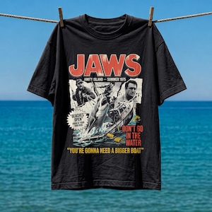 Könnte beinhalten: Ein schwarzes Kurzarm-T-Shirt mit einem Grafikdruck zum Film Jaws. Das Design zeigt den Titel JAWS, Bilder von Charakteren, einen Hai, ein Boot sowie den Text Beaches Open, Don't Go In The Water und You're Gonna Need A Bigger Boat.