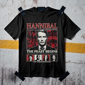 Könnte beinhalten: Ein schwarzes Kurzarm-T-Shirt mit einem Grafikdruck, der den Text HANNIBAL, THE FEAST BEGINS und A BEAUTIFUL MIND CAN BE A DANGEROUS PLACE enthält. Das Design zeigt ein zentrales Porträt eines Mannes im Anzug, umgeben von kleineren Charakterbildern.