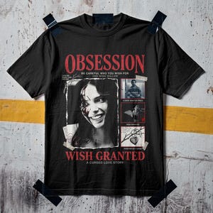 May include: A black short-sleeve t-shirt featuring a graphic print with the text OBSESSION, WISH GRANTED, and A CURSED LOVE STORY. The design includes a black and white portrait of a smiling person with blood on their face and three smaller panels.