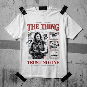 Puede incluir: Una camiseta blanca de manga corta con un estampado gráfico que incluye el texto THE THING, OUTPOST 31 - ANTARCTICA, WINTER 1982, TRUST NO ONE y ASSIMILATION IS SURVIVAL. El diseño muestra una figura barbuda, un perro e imágenes científicas.