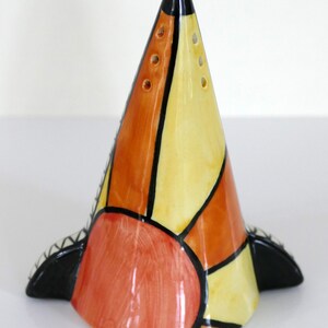May include: A conical ceramic shaker featuring a segmented pattern of yellow, orange, and red sections outlined in thick black lines. The top of the cone has small holes, and the base includes two black, wing-like protrusions on either side.