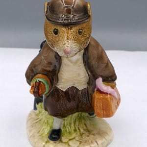 May include: A ceramic figurine of a mouse dressed in a brown jacket, a matching cap, and a white waistcoat. The mouse holds a small orange suitcase with a pink handle in one hand and a green-handled umbrella in the other, standing on a base.