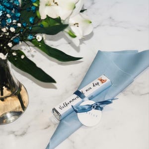 May include: A light blue folded napkin with a small gift box and a round tag tied with a blue ribbon. The gift box has text that reads "Velkommen i min dåp 2026". A vase of white lilies and blue flowers is in the background.