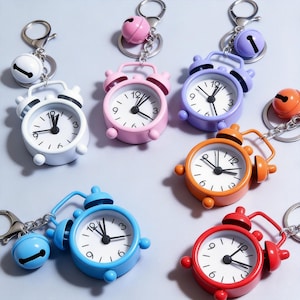 May include: A collection of six miniature alarm clock keychains in white, pink, purple, orange, blue, and red. Each keychain features a small round clock face with black numbers and hands, a metal ring, and a matching colored jingle bell charm.