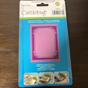 May include: A Cuttlebug Grace's Frame embossing folder in original retail packaging. The blue card backing features the product name, a barcode, and three instructional images showing the folder being used with a die-cutting machine.