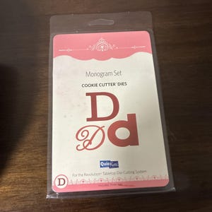 May include: A QuicKutz Monogram Set Cookie Cutter Dies package for the Revolution Tabletop Die-Cutting System. The packaging is white and pink with the letter D displayed in three different fonts, including a serif, sans-serif, and script style.