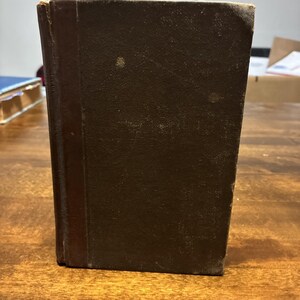 Könnte beinhalten: Ein altes gebundenes Buch mit einem dunkelbraunen, strukturierten Einband und einem vertikalen Buchrückenstreifen. Das Buch ist rechteckig und weist leichte Gebrauchsspuren an den Kanten und Ecken des Vorderdeckels sowie eine glatte Oberfläche auf.