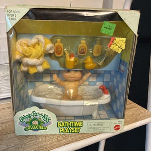 May include: A Cabbage Patch Kids Bathtime Playset in original packaging. The set includes a small doll in a white bathtub with molded bubbles, a yellow towel, three yellow bottles, a yellow mug with a toothbrush, and a yellow rubber duck.