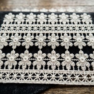 Peut inclure: Une serviette gris anthracite foncé ornée d'un large panneau rectangulaire de dentelle blanc cassé. La dentelle présente un motif répétitif de formes florales disposées en rangées horizontales, avec une bordure délicatement festonnée.