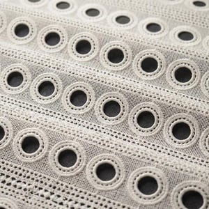 May include: A cream-colored fabric featuring a repeating pattern of embroidered circular eyelets arranged in horizontal rows. The material has a semi-sheer, mesh-like texture with stitched borders surrounding each circular opening.