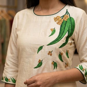 May include: An off-white long-sleeved top featuring a round neckline with dark green trim. The garment is decorated with embroidered green eucalyptus leaves and small yellow flower clusters across the chest and on the sleeve cuffs.