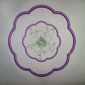 May include: A circular embroidered patch featuring a scalloped purple border. The outer ring has a light gray background with a white geometric pattern. The center is a solid light gray circle with a green floral design and a small pink center.