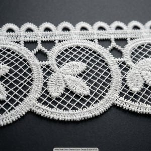 May include: A white cotton chemical lace trim featuring a repeating pattern of circular motifs with internal grid detailing and leaf-like embroidery. The top edge has a scalloped border, and the bottom edge features a continuous scalloped design.