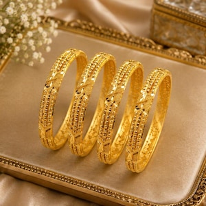 May include: Four gold bangles featuring a textured surface with repeating geometric patterns and beaded detailing. Each circular bracelet has a polished finish and a consistent width, showcasing intricate metalwork across the entire outer band.