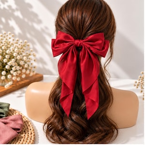 May include: A large, deep red fabric hair bow with long, flowing tails secured in the center of brown, wavy hair. The bow features a smooth, satin-like texture and is positioned at the back of the head, cascading down over the hair strands.
