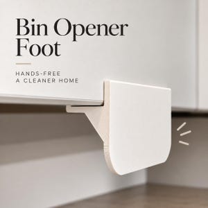 Pode incluir: Um abridor de lixeira de pé branco fixado a uma porta de armário, com o texto Bin Opener Foot e HANDS-FREE A CLEANER HOME. O dispositivo possui um painel plano retangular com cantos inferiores arredondados e um suporte na borda.