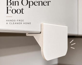 May include: A white bin opener foot attached to a cabinet door, featuring the text Bin Opener Foot and HANDS-FREE A CLEANER HOME. The device has a flat, rectangular panel with rounded bottom corners and a support bracket extending to the cabinet edge.