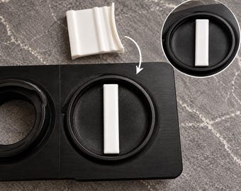 May include: A black rectangular base with two circular recessed compartments. One compartment contains a vertical white rectangular insert. A separate white curved piece is shown above the base, with an arrow indicating placement.