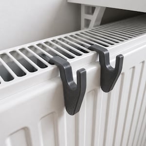 Puede incluir: Dos ganchos de plástico gris oscuro diseñados para colgarse sobre el borde superior de un radiador de metal blanco. Cada gancho presenta un perfil curvo en forma de J que encaja de forma segura en la rejilla superior del radiador.