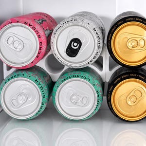 Pode incluir: Seis latas de bebida de alumínio dispostas numa grelha de dois por três dentro de um suporte de plástico branco. A fila superior apresenta uma lata cor-de-rosa, uma branca com texto preto e uma preta com topo dourado. A fila inferior tem duas latas verde-azuladas e uma preta.