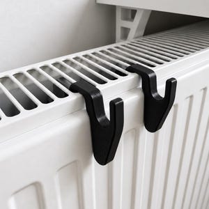 Può includere: Due ganci neri a forma di J progettati per essere appesi al bordo superiore di un termosifone bianco. I ganci presentano una sezione superiore curva che poggia sulle alette del radiatore e un braccio verticale rivolto verso il basso.