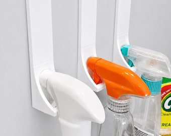 May include: Three white over-the-cabinet hooks made of rigid plastic, each featuring a flat top bracket and a curved lower section designed to hold the neck of a spray bottle. The hooks are mounted side-by-side on a flat, light-colored surface.