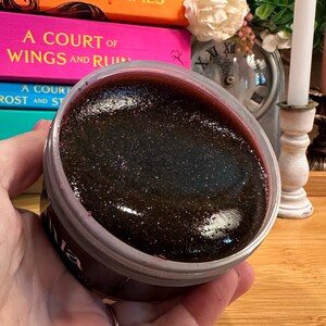 May include: A jar of dark, shimmering body scrub with a deep red rim. The product is held in a clear plastic container. The background includes stacked books with titles like "A Court of Silver Flames" and a lit candle.