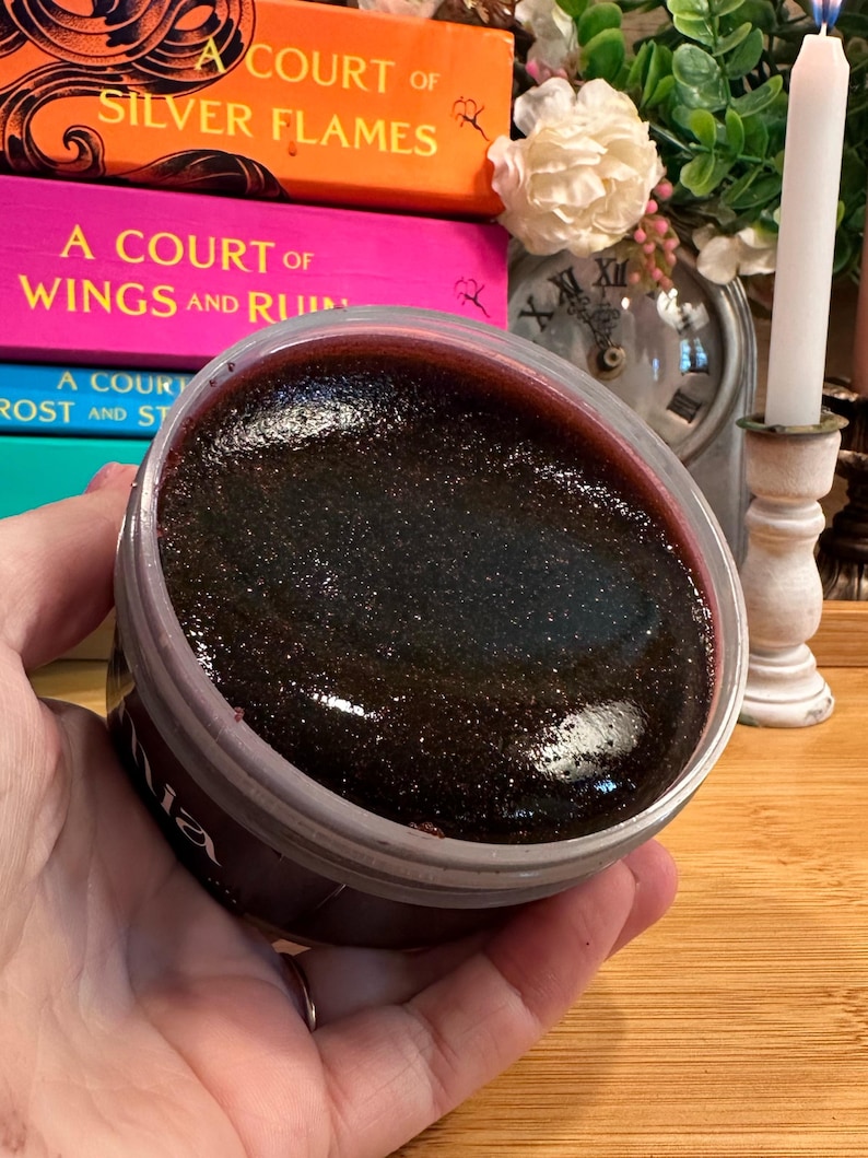 May include: A jar of dark, shimmering body scrub with a deep red rim. The product is held in a clear plastic container. The background includes stacked books with titles like "A Court of Silver Flames" and a lit candle.