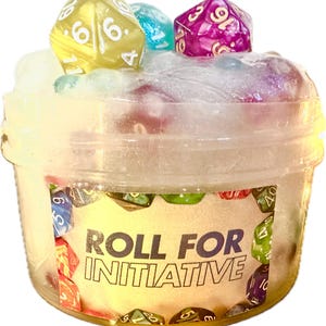 May include: A clear plastic container filled with a white, fluffy substance, topped with colorful dice. The container has a label that reads "ROLL FOR INITIATIVE". The dice are yellow, blue, and purple.