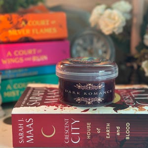Puede incluir: Una vela encendida en un portavelas de madera junto a una vela en tarro con la etiqueta "Dark Romance" sobre un libro. Otros libros est&aacute;n apilados en el fondo, con t&iacute;tulos como "A Court of Silver Flames".