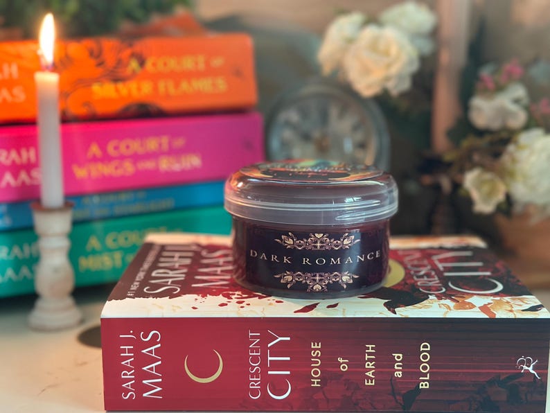Puede incluir: Una vela encendida en un portavelas de madera junto a una vela en tarro con la etiqueta "Dark Romance" sobre un libro. Otros libros est&aacute;n apilados en el fondo, con t&iacute;tulos como "A Court of Silver Flames".