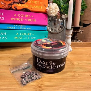 May include: A stack of colorful books with titles like "A Court of Silver Flames" and "A Court of Wings and Ruin" sits next to a candle, a small clock, and a container labeled "Dark Academia". A small bag of miniature books is also present.