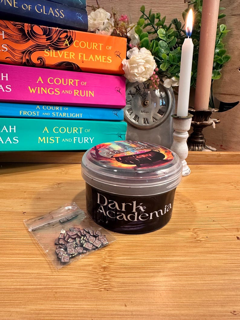 May include: A stack of colorful books with titles like "A Court of Silver Flames" and "A Court of Wings and Ruin" sits next to a candle, a small clock, and a container labeled "Dark Academia". A small bag of miniature books is also present.