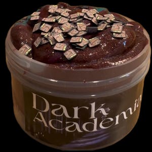 May include: A jar of dark brown slime with glitter and small, square, patterned decorations on top. The jar has a clear lid and a label that reads "Dark Academia" in a stylized font.