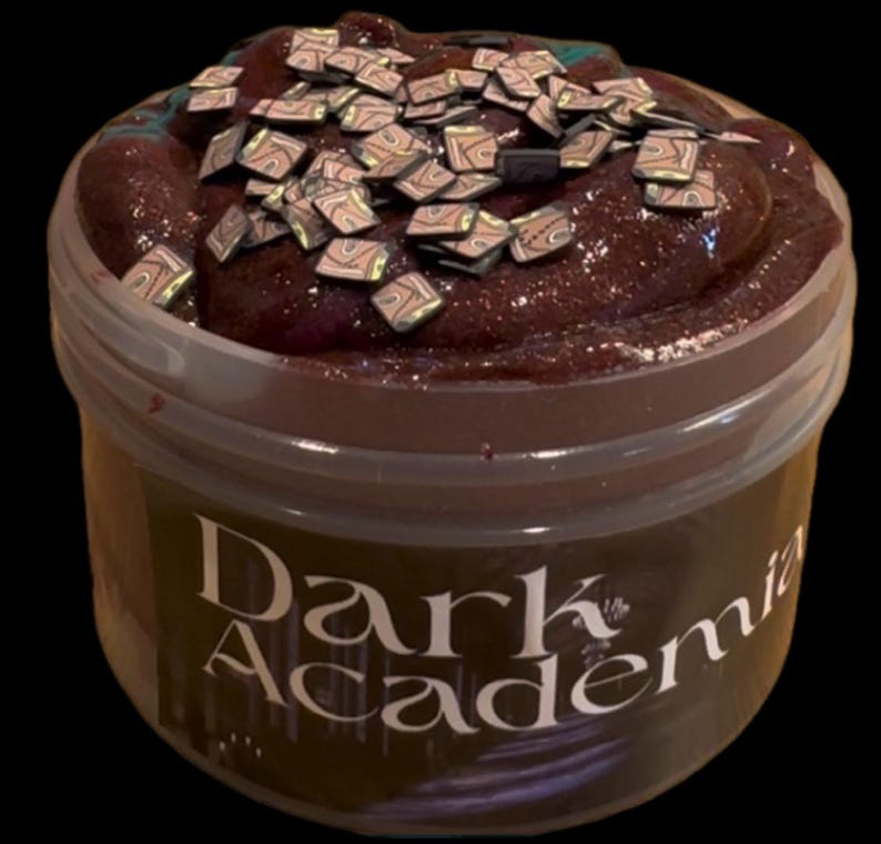 May include: A jar of dark brown slime with glitter and small, square, patterned decorations on top. The jar has a clear lid and a label that reads "Dark Academia" in a stylized font.