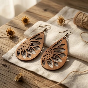 May include: A pair of teardrop-shaped wooden earrings featuring a laser-cut sunflower design. The earrings have dark brown metal hooks and a natural wood grain finish, showcasing intricate petal cutouts and a central seed pattern.