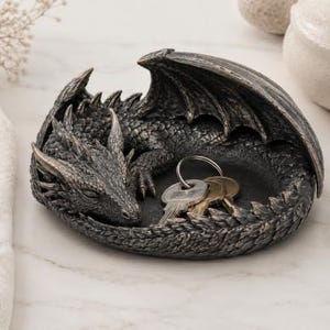 Può includere: Una statuetta di drago con finitura metallica grigio scuro, modellata a forma di ciotola o vassoio poco profondo. Il drago è raffigurato in posizione rannicchiata e a riposo, con le ali ripiegate sul dorso. Un portachiavi con diverse chiavi poggia all'interno.