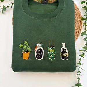 Può includere: Una felpa girocollo verde bosco con quattro ricami: un gatto nero in un vaso arancione con una pianta, un gatto nero dentro un barattolo di vetro, un gatto nero in una pianta sospesa e un gatto nero dentro una bottiglia con dello spago giallo.