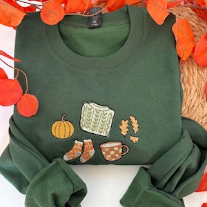 Peut inclure: Un sweat-shirt à col rond vert forêt foncé avec un motif brodé sur la poitrine. La broderie représente une citrouille jaune, un pull en tricot torsadé vert clair, une paire de chaussettes à motifs, une tasse marron et des feuilles de chêne orange.