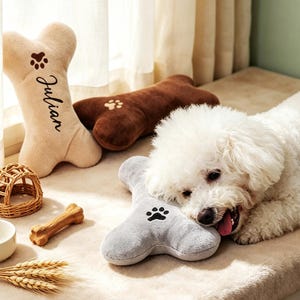 May include: A white curly-haired dog resting its head on a grey bone-shaped plush toy. Nearby are a beige bone-shaped plush toy with the name Julian and a paw print, a brown bone-shaped plush toy, a small wicker dome, a bone chew, and wheat stalks.