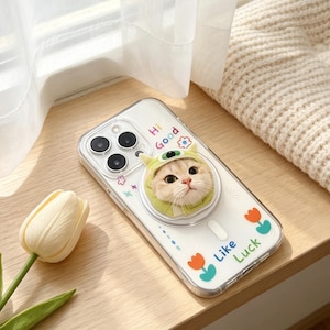 May include: A clear smartphone case featuring a circular pop-out grip with a plush cat wearing a green monster hood. The case is decorated with colorful small flower and star icons, along with the printed text "Hi Good" and "Like Luck".