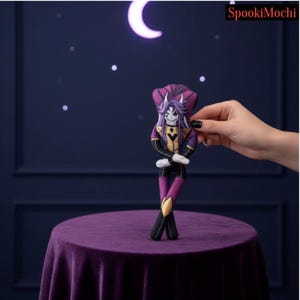 Puede incluir: Una muñeca de peluche plana que representa un personaje con cabello morado, cuernos blancos y un atuendo negro y dorado. El personaje tiene una barbilla puntiaguda, una sonrisa amplia y los brazos cruzados. El texto SpookiMochi aparece arriba.