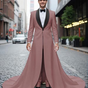 May include: A dusty rose long-line tuxedo jacket featuring a black shawl lapel, a single front button, and a floor-length train. The jacket is worn over a crisp white button-down shirt with a black bow tie and matching dusty rose trousers.
