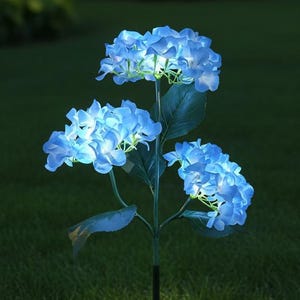 以下が含まれることがあります： 3つの青く光る花房を持つアジサイの形をしたソーラーガーデンライト。この人工植物には緑の葉があり、細い黒い金属製の支柱が地面に差し込まれており、芝生を照らしています。