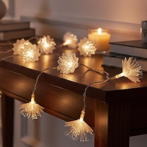 May include: A string of decorative LED lights featuring translucent, flower-shaped covers. Each light emits a warm, soft glow, with the individual blossoms arranged along a thin, flexible wire that is draped across a dark wooden surface.