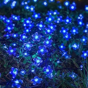 Op de afbeelding: Een snoer met decoratieve bloemvormige led-lampjes die een helderblauw licht uitstralen, verspreid over groen gebladerte. Elk individueel lampje heeft vijf bloemblaadjes en is verbonden door een dunne donkere draad voor een lichtgevend effect.