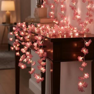 Op de afbeelding: Een decoratieve lichtsnoer met kleine roze kersenbloesems. De bloemenslinger is gedrapeerd over een donkerhouten sidetable, waarbij de individuele bloesems worden verlicht door warmwitte LED-lampjes die over de gehele lengte zijn verspreid.