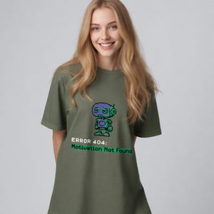Könnte beinhalten: Ein olivgrünes Kurzarm-T-Shirt mit einer Pixel-Art-Grafik eines violetten und grünen Roboters mit müdem Gesichtsausdruck. Unter dem Roboter steht der Text "ERROR 404: Motivation Not Found" in weißer und grüner, verpixelter Schrift.